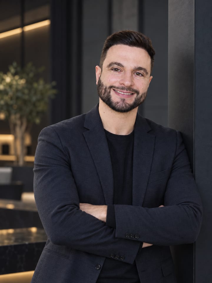 Luiz Hogrefe, smiling, arms crossed, in a dark blazer over a black knit top, leaning against a dark wall